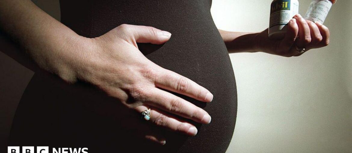 Stock image: Pregnant woman with dark top holding her stomach with her right hand and wearing a ring. In her left hand she is holding two vitamin containers.