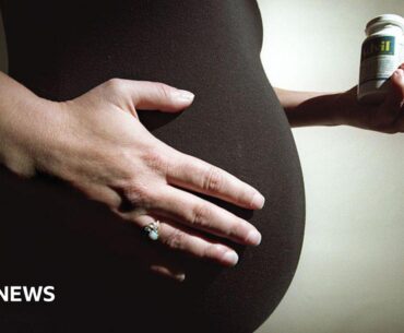Stock image: Pregnant woman with dark top holding her stomach with her right hand and wearing a ring. In her left hand she is holding two vitamin containers.