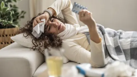 Getty Images Sick young woman lying on the couch holding a blister pack with antibiotics. 