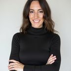 A woman with wavy brown hair and brown eyes, wearing a black turtleneck, smiles while holding her arms crossed.