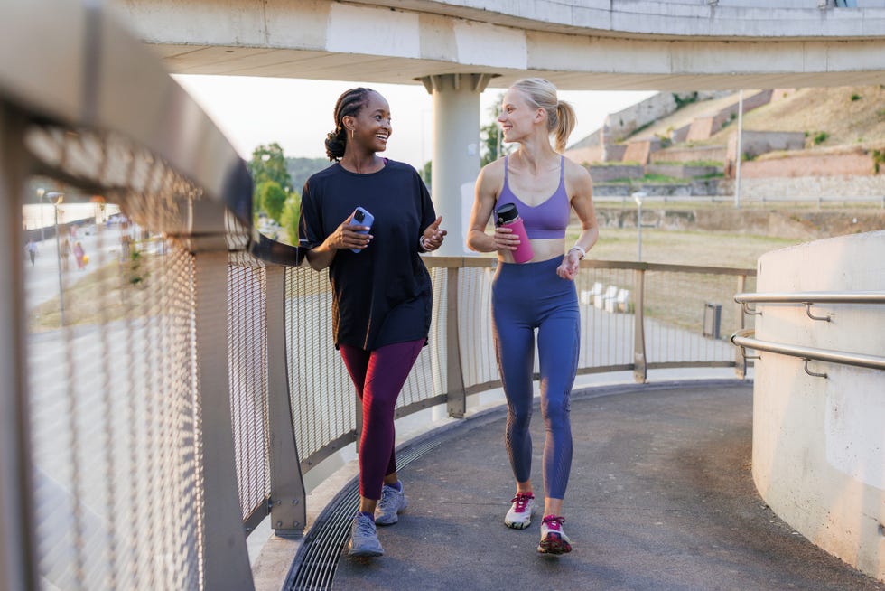 Two diverse young women on a power walk outdoors two friends are jogging together on a raised outdoor pathway, engaging in conversation. they appear energetic and relaxed, enjoying their fitness routine and the pleasant surroundings.