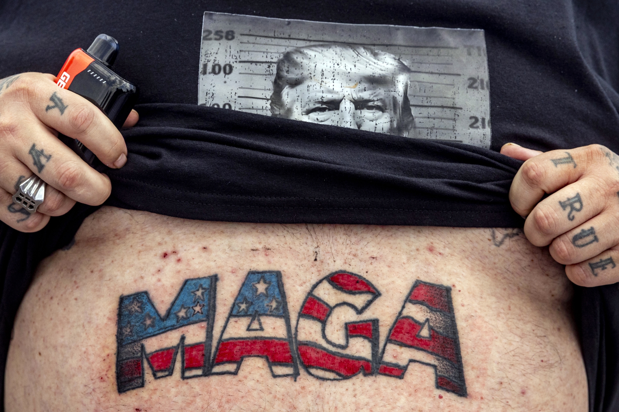 Casey Boeling, 46 of Livonia, shows off a brand new MAGA tattoo on his stomach while pulling up his T-shirt that shows President Donald J. Trump in a mugshot as hundreds of supporters wait in line to celebrate Trump, where he will deliver as speech about the successes of his first 100 days in office on Tuesday, April 29, 2025 at Macomb Community College in Warren.