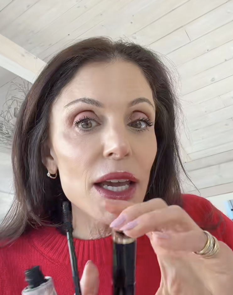 A woman in a red sweater holds up a mascara wand and tube.