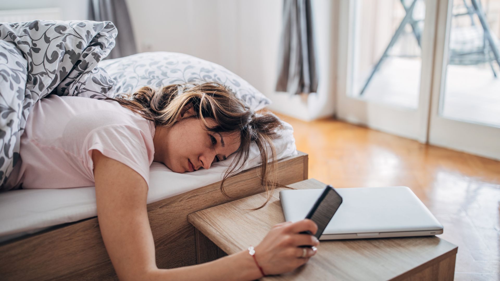 A woman lies on her front in bed looking tired as she holds her phone in front of her while resting her hand on the bedside table.