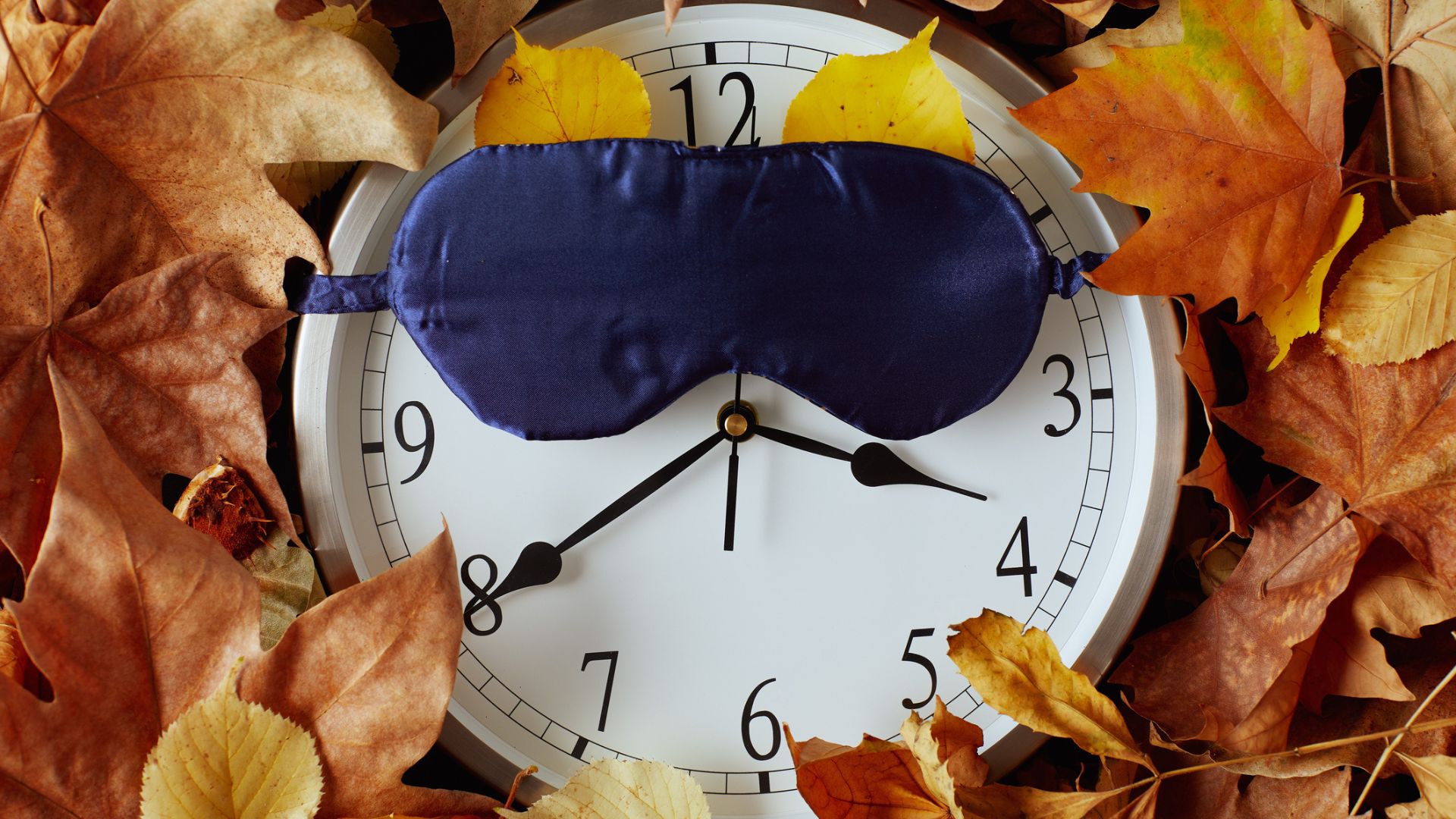 An image of an alarm clock buried in autumnal leaves with a blue silk sleep mask pulled over it
