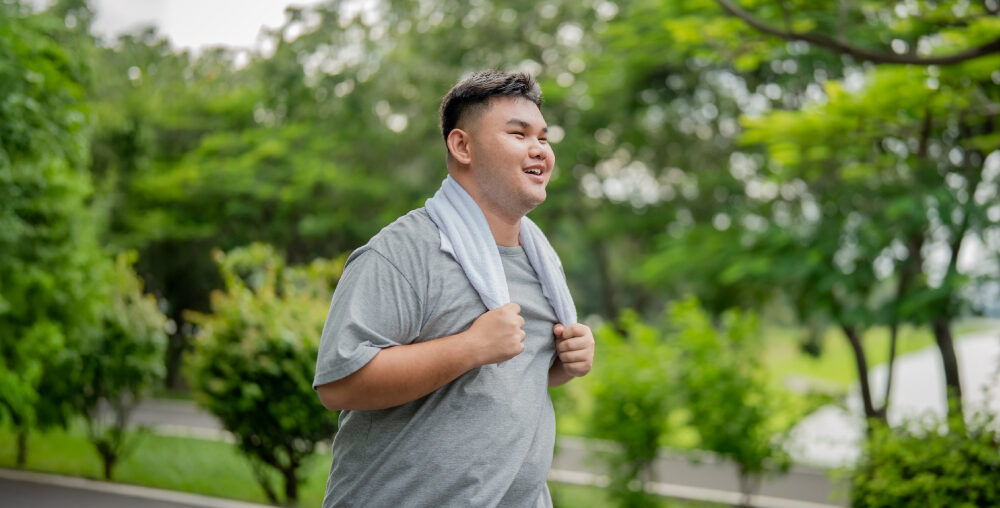 A smiling man is seen walking outdoors in a lush green park setting. He is wearing a gray shirt and has a towel draped around his neck. The background features trees and a pathway, creating a serene and natural environment.