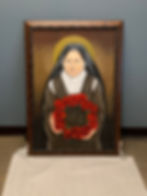 A framed painting of a nun with a halo holding a wreath of red roses, set against a plain gray wall. The mood is serene and reverent.