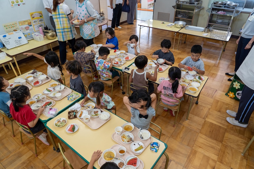 Children sit at various tables eating food in a Japanese childcare centre.