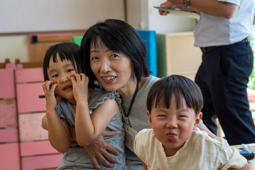 A Japanese woman smiles with her arms around her daughter and son as they both pull funny faces.