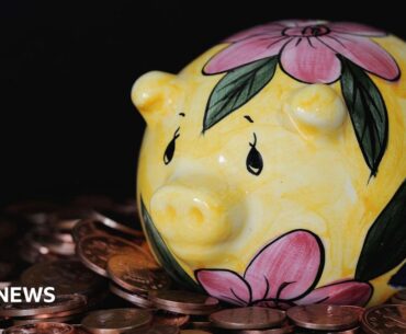 The image shows a yellow ceramic piggy bank with pink flowers and green leaves painted on it. It has black eyes and it is resting on a pile of coins.
