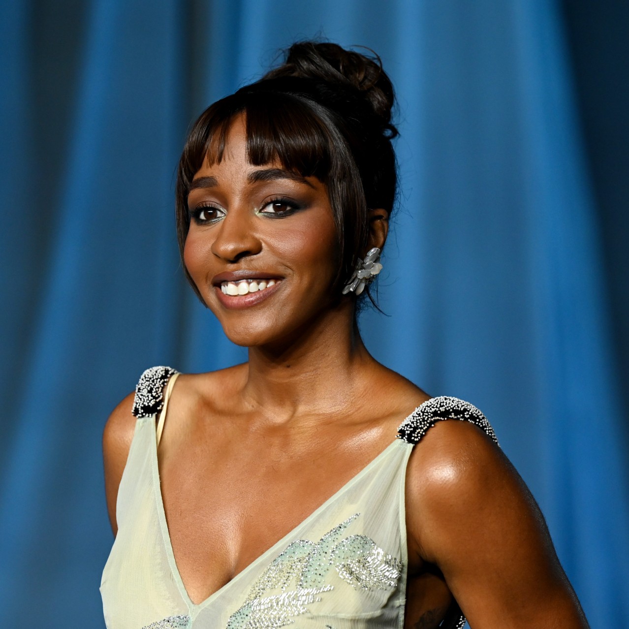 Ayo Edebiri with micro bangs and dark hair posing on a red carpet in front of a blue background.
