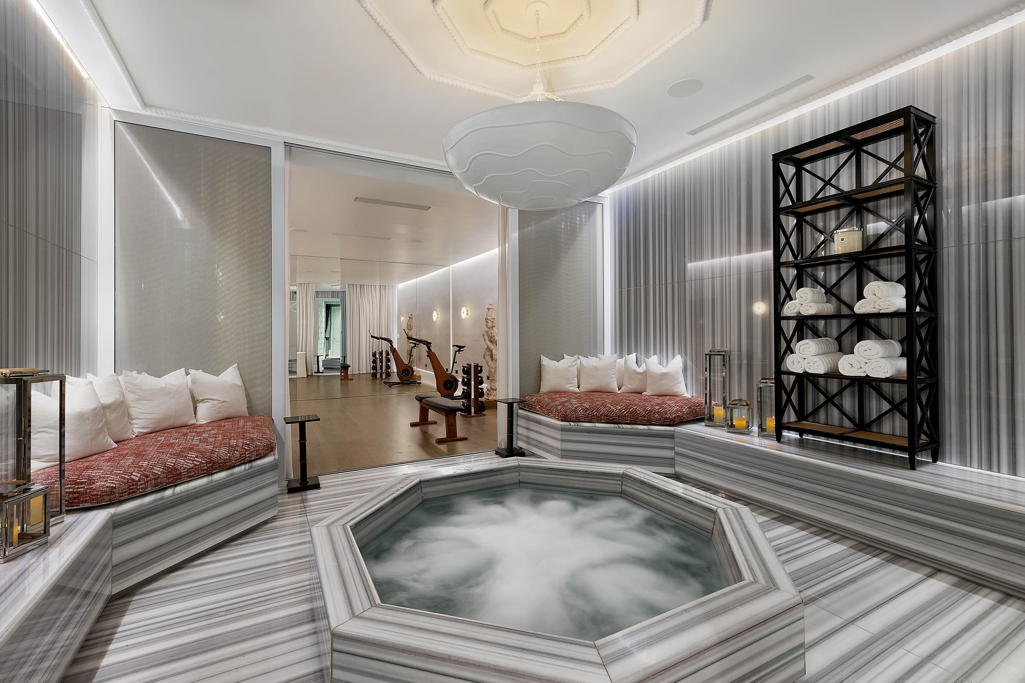 A luxurious meditation room with a hexagonal hot tub, marble floors, sitting areas with pillows, a black shelving unit with rolled towels, and a view into a mirrored gym.