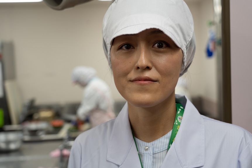 A Japanese woman smiles while wearing a chef's hat and uniform