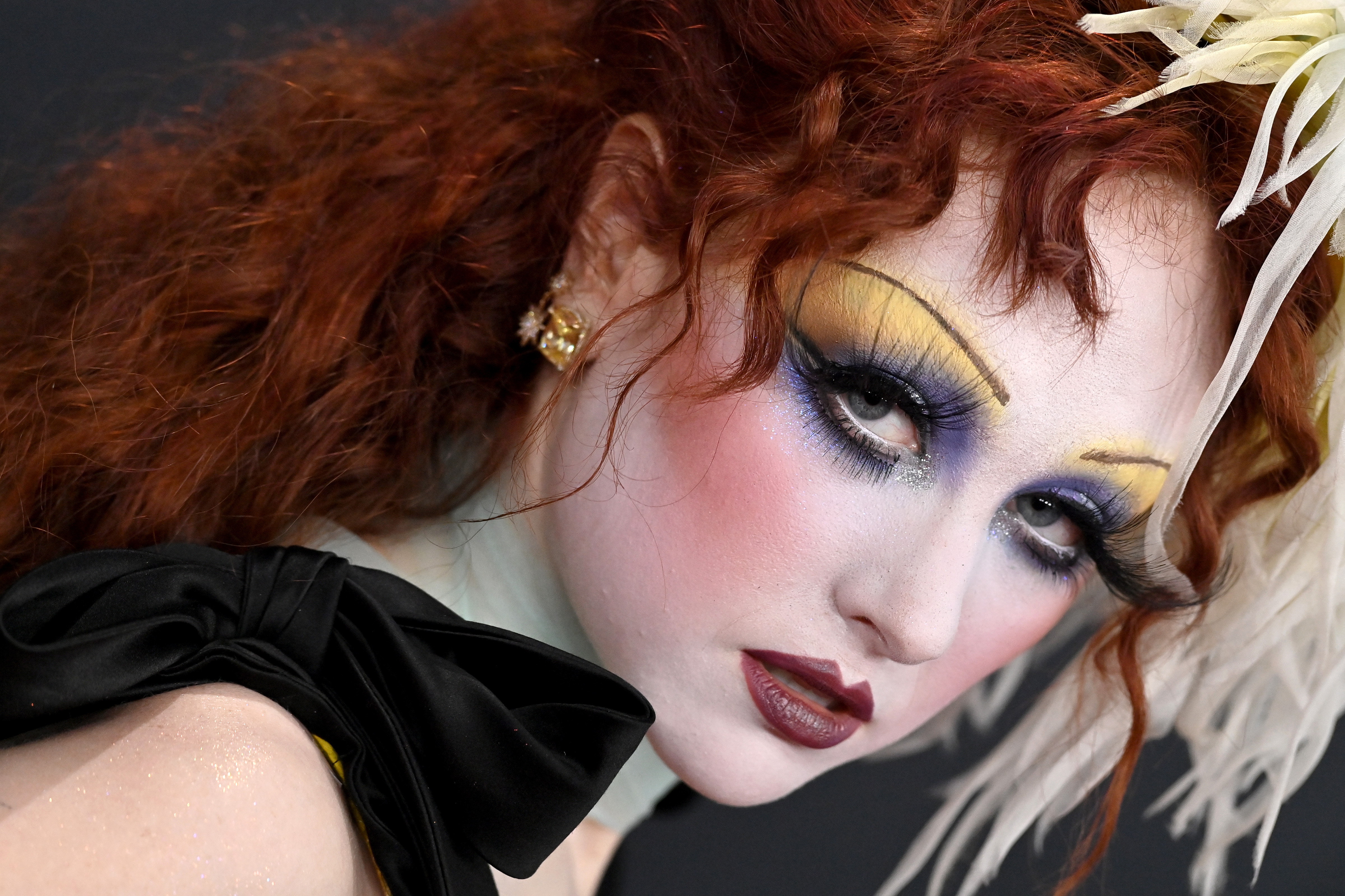 Chappell Roan attends the 67th GRAMMY Awards with dramatic makeup including yellow and purple eyeshadow, and dark red lips, complemented by red curly hair and a black bow on her shoulder.