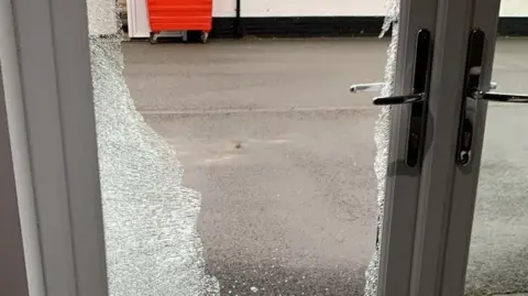 M & Arc Hair and Beauty A shattered white-framed glass door with silver handles. It has a broken glass panel inside it.