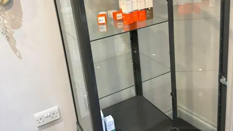 M & Arc Hair and Beauty An empty glass cabinet with only a few orange and white boxes of products left on the top and bottom shelves.