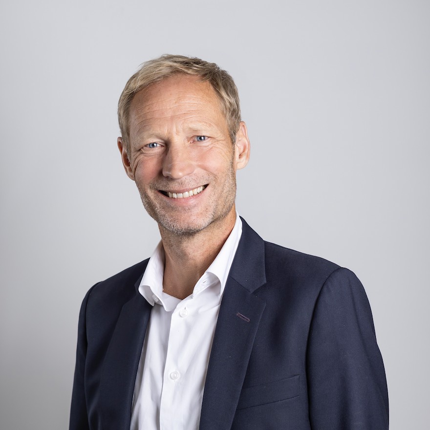 A man wearing a white shirt and dark blue jacket smiles at the camera.