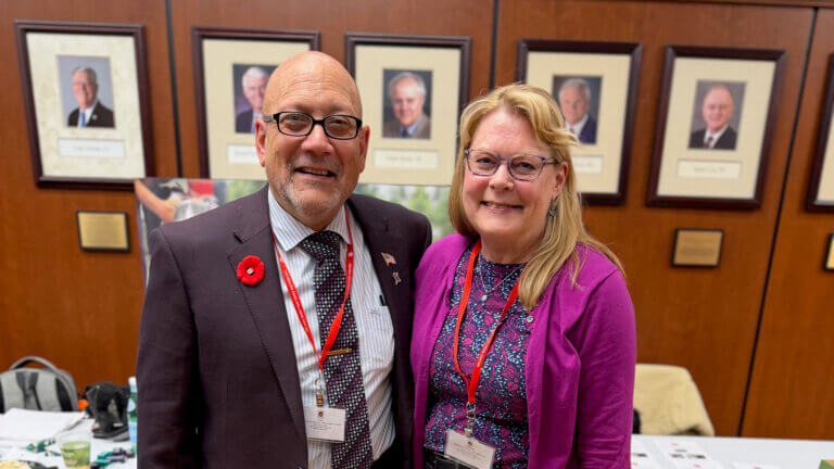 two people standing in front of a wall featuring awards. The man on the left is wearing a jacket with a red floral button. The woman on the right is wearing a purple shirt and jacket and has long, blonde hair.