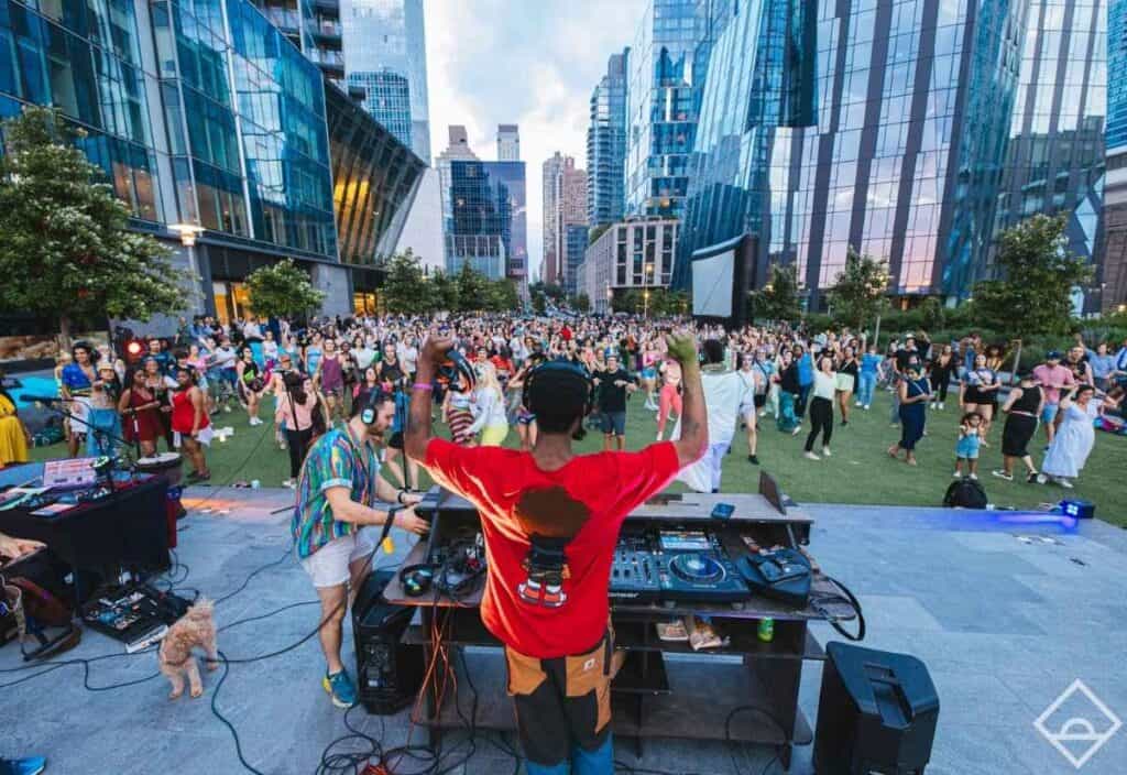 A DJ plays music on an outdoor stage as a large crowd gathers on a grassy plaza surrounded by modern glass buildings in the city.