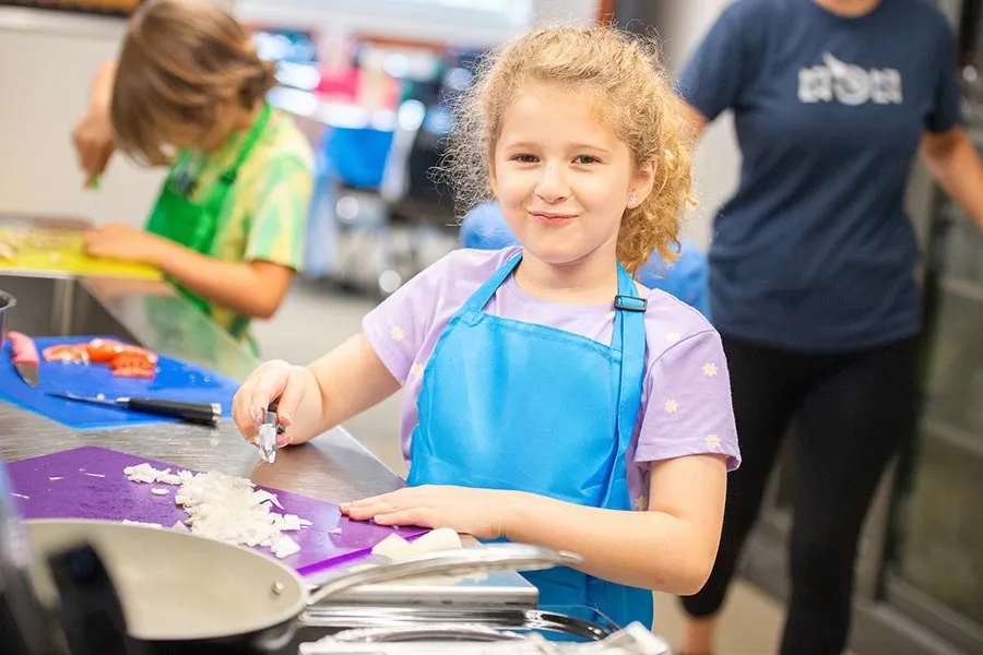 a young girl in a blue apron smiles at camera while cutting onions