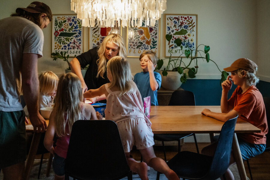 A group of children and two adults gather around a dining table with snacks and games under a chandelier in a brightly decorated room, enjoying laughter and fun on Jasmin Day.