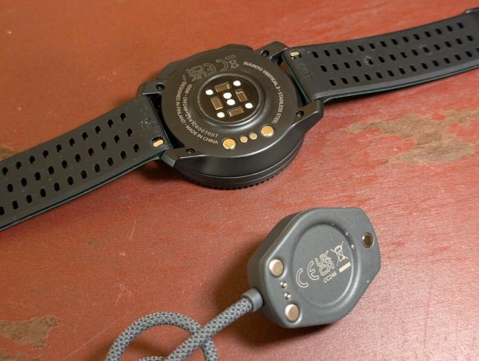 Close-up of the underside of a black smartwatch and its charging dock, featuring visible sensors and branding.