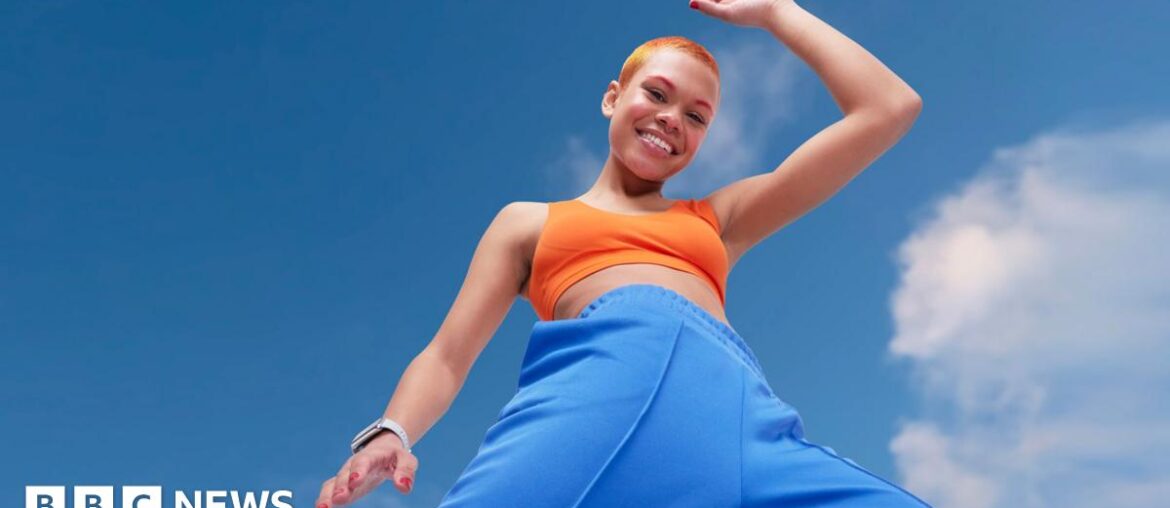 Young woman looking energetic and vibrant. She has blue tracksuit bottoms and orange crop top and orange hair. She is striding energetically in front of a blue sky