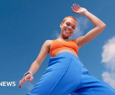 Young woman looking energetic and vibrant. She has blue tracksuit bottoms and orange crop top and orange hair. She is striding energetically in front of a blue sky