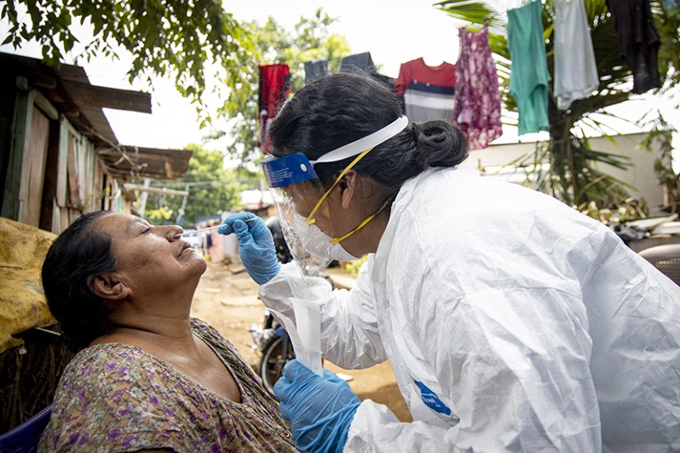 On the left, a woman sits with her head back and eyes closed. On the right, a woman wearing white coat, blue gloves, and face mask, takes nasal swab from the seated woman.