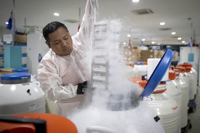 A man wearing a white coat is holding a piece of equipment in an open storage container. A steam-like gas emanates from the container. He is surrounded by other similar containers in the room.