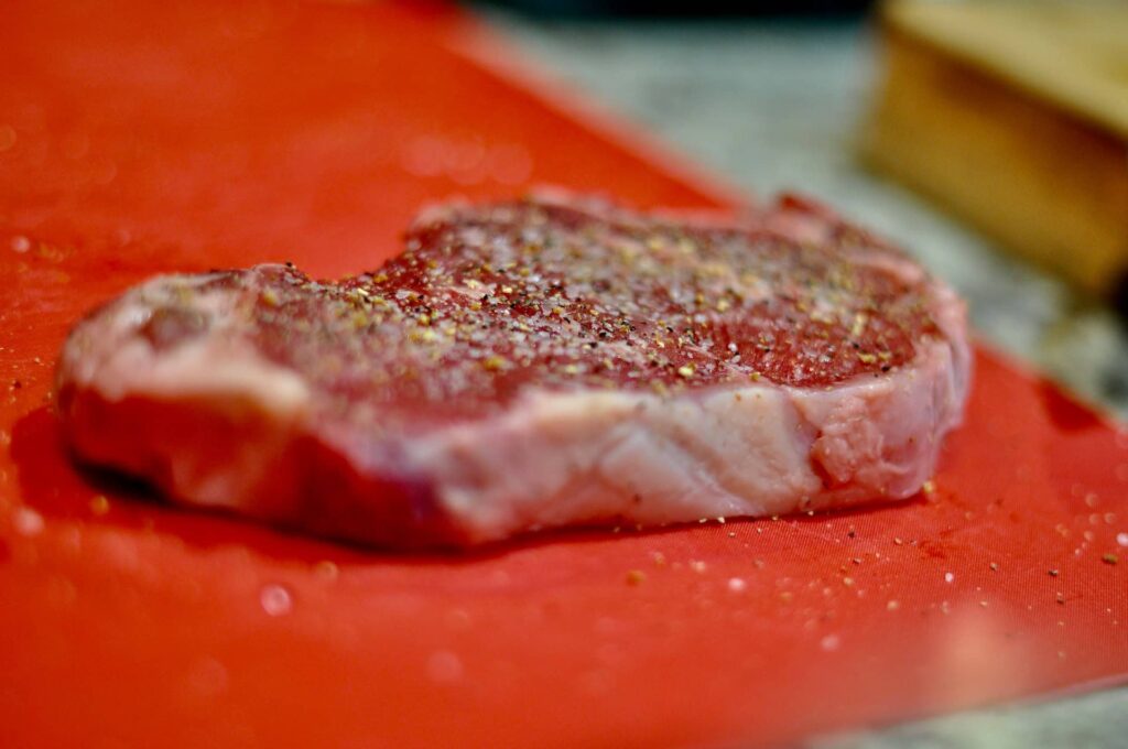 Raw steak seasoned with salt and black pepper on a red cutting board.