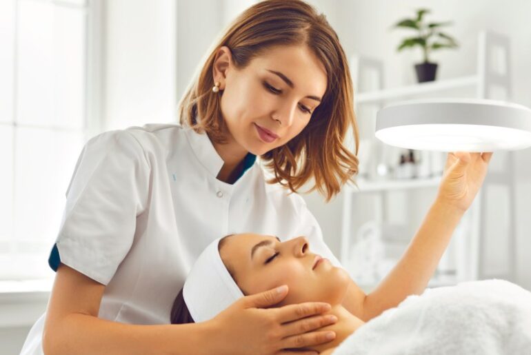 A cosmetologist or dermatologist directing lamp for facial treatment to young woman.