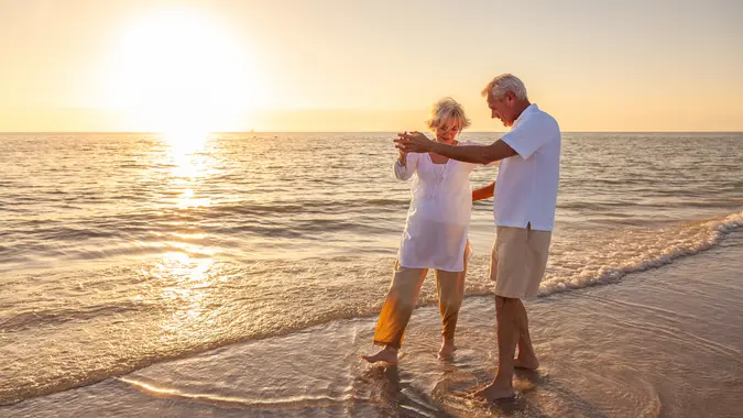 Happy senior man and woman old retired couple walking dancing and holding hands on a beach at sunset.