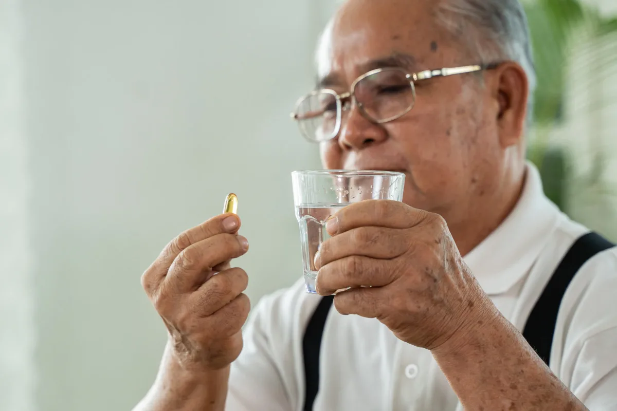 Senior Asian man with cup of water and pill looking at camera and taking remedy in cozy room at home