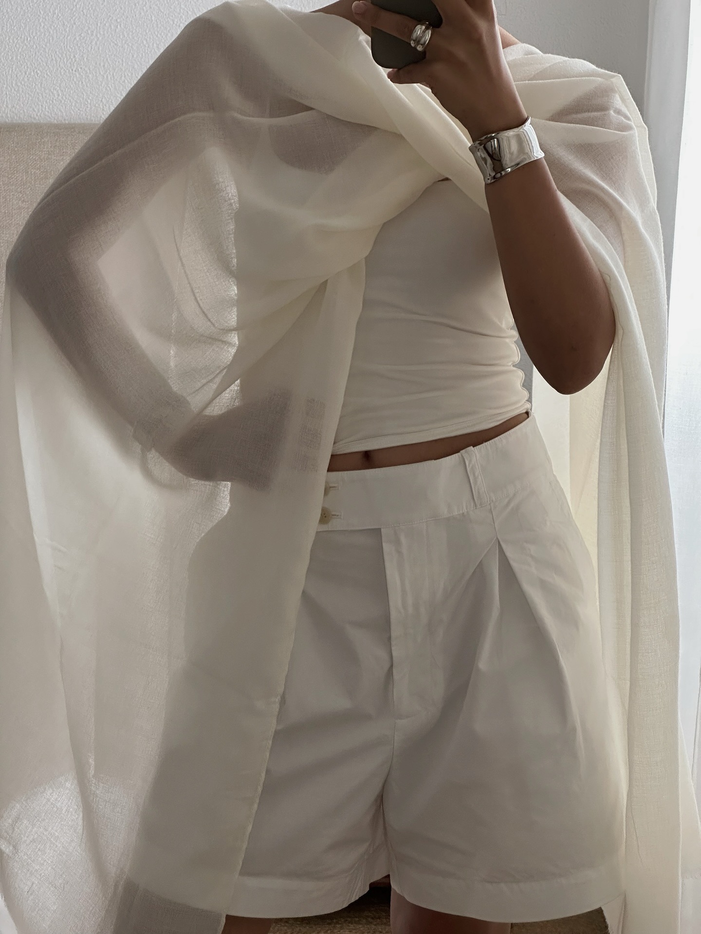 A woman taking a mirror selfie in a crisp white top, shorts and sheer white cape
