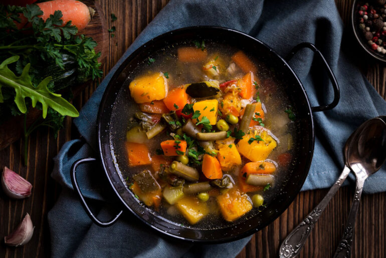 A hearty, warming vegetable soup in a black bowl on a wooden table, with vibrant carrots, peas, and squash. Fresh herbs, garlic, and spoons add a rustic touch.