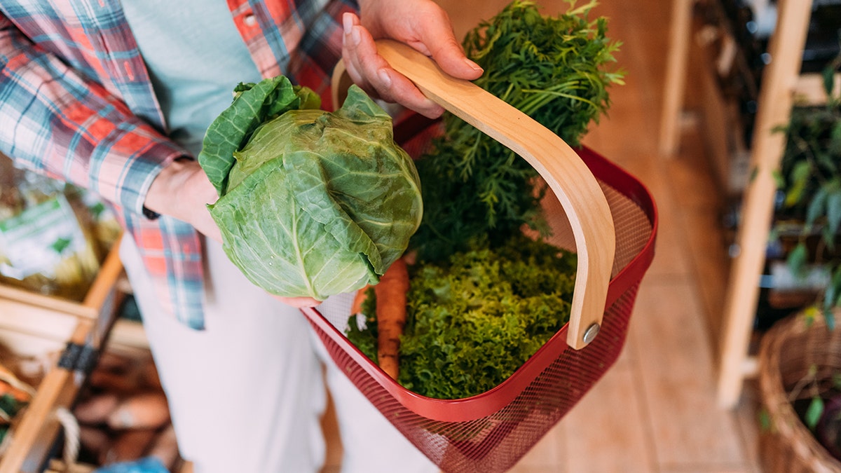 Person holding a green cabbage and a red shopping basket filled with fresh carrots, leafy greens, and herbs at a local grocery store.