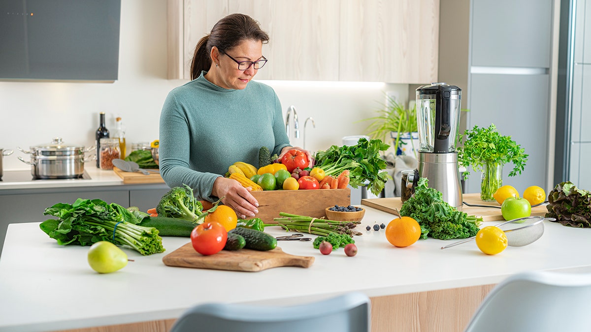 Woman preparing fresh fruits and vegetables on a kitchen counter with a blender, focusing on healthy home cooking.
