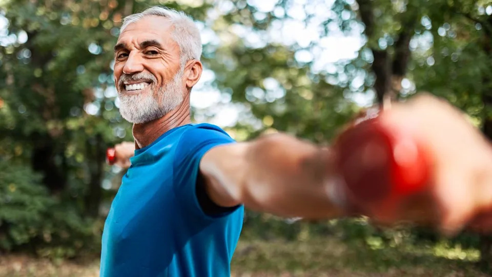  Older male in blue t-shirt holding dumbbells out at shoulder-height in a local park smiling. 