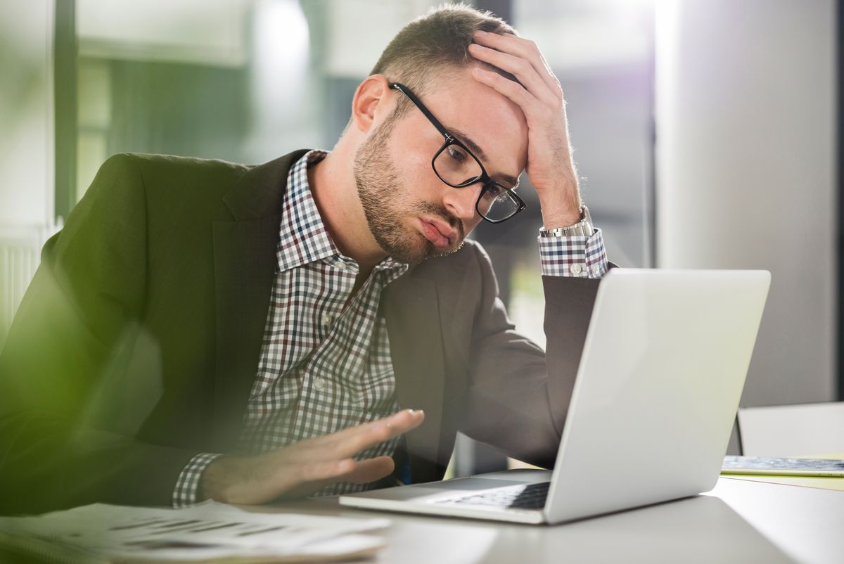 Man looks stressed holding his head in his hands as he sits at desk looking at laptop