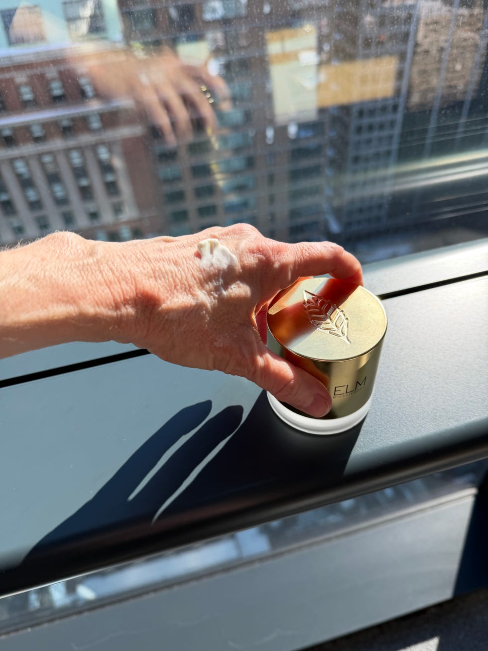 A hand holding a container of skincare product next to a window. A hand holding a container of skincare product next to a window.