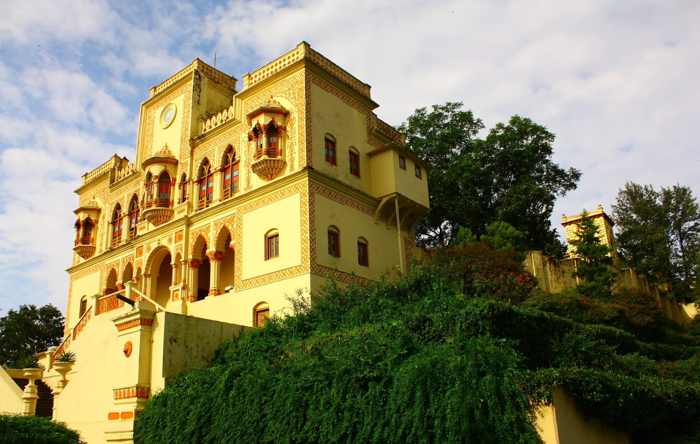 Ananda in the Himalayas building, a large yellow structure with intricate red and orange patterns, stands on a hill surrounded by lush green foliage.