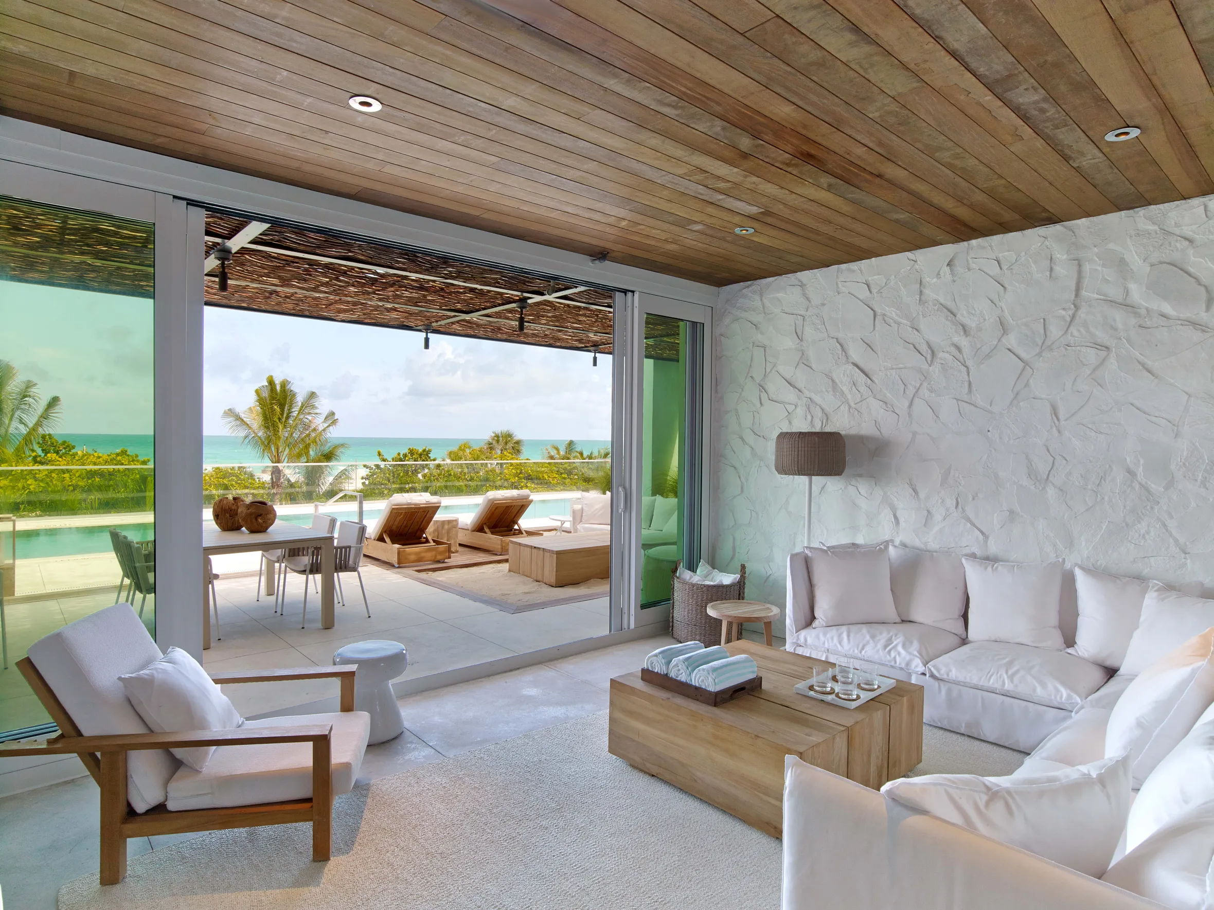 Living room with white couches and wood furniture, opening onto a patio with lounge chairs overlooking a beach and ocean.