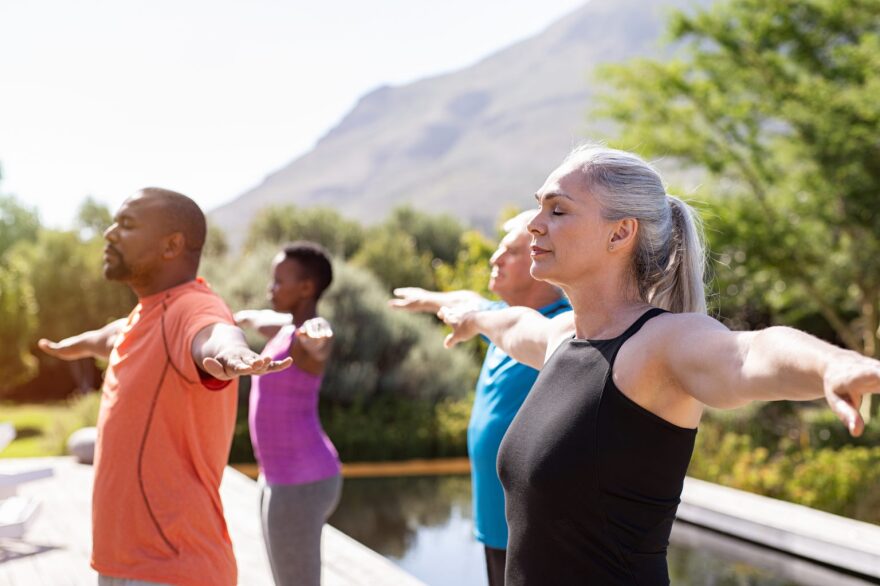 Group of senior people with closed eyes stretching arms outdoor. Happy mature people doing breathing exercise near pool. Yoga class with women and men doing breath exercise with outstretched arms. Balance and meditation concept. (Group of senior peopl