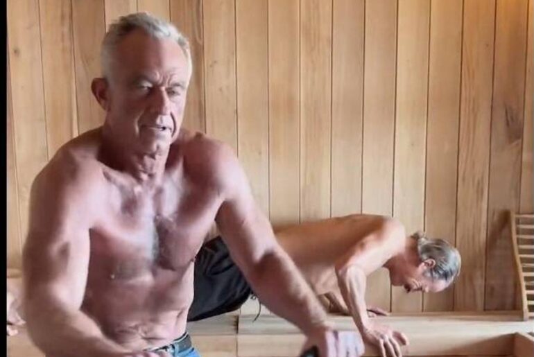 An elderly man is seated in a wooden-walled room, possibly in a sauna, with his arms resting on his knees. Behind him, another individual is performing push-ups on the floor.