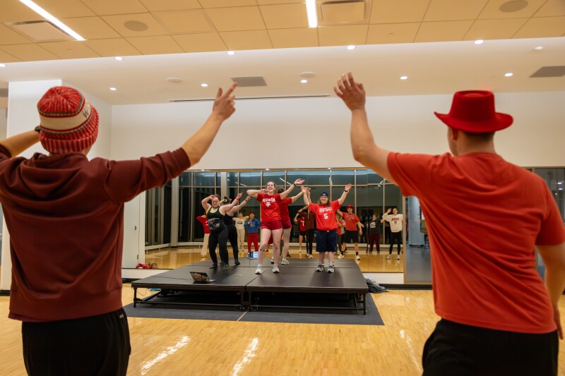 Three people on an elevated platform lead a fitness class as others in the room follow along.