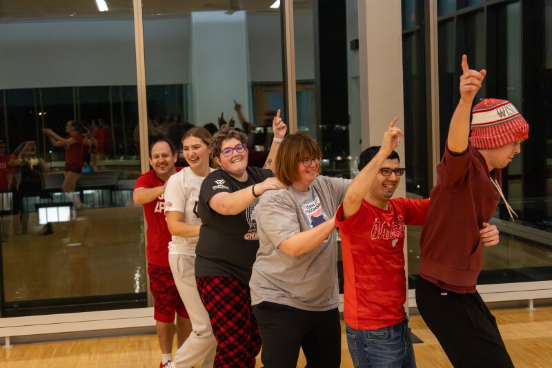 A group of people stand in a conga line with their hands on each other’s shoulders.
