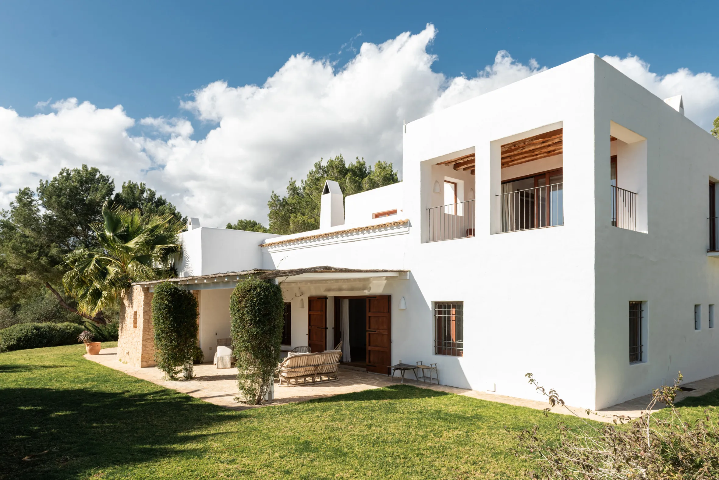 White, multi-level house with balconies, a patio, and a grassy lawn, surrounded by trees.