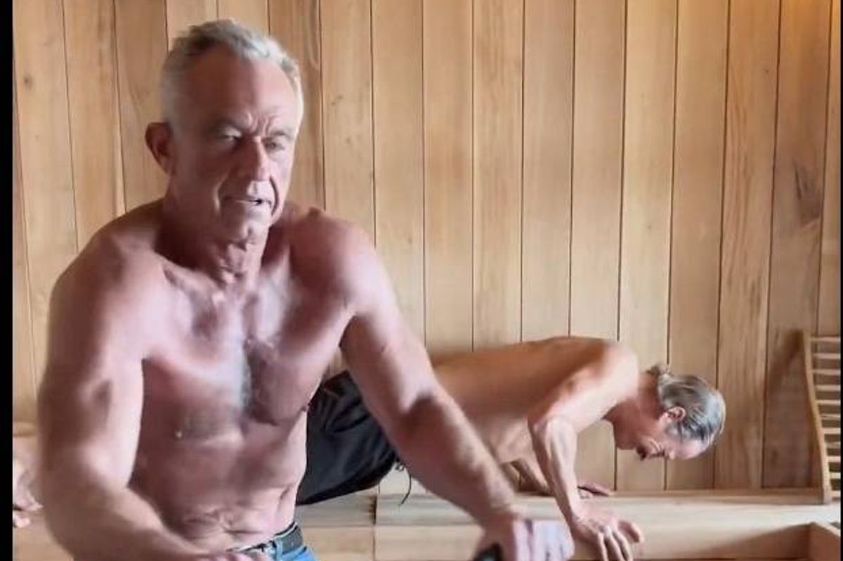 An elderly man is seated in a wooden-walled room, possibly in a sauna, with his arms resting on his knees. Behind him, another individual is performing push-ups on the floor.