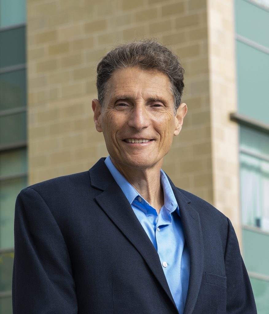 Portrait photo of Mitchell Kronenberg. He is wearing a suit and smiling at the camera. He is standing outside, in front of the LJI building.